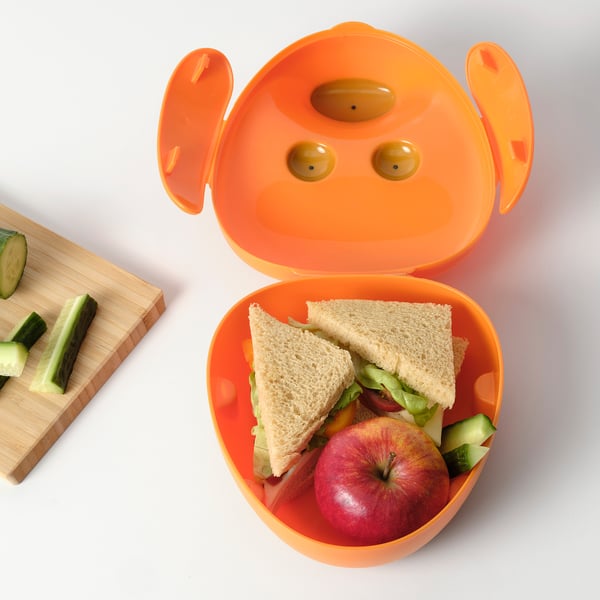 Bright orange SMASKA container with ears open, holding a sandwich and apple next to a wooden cutting board with cucumber slices.