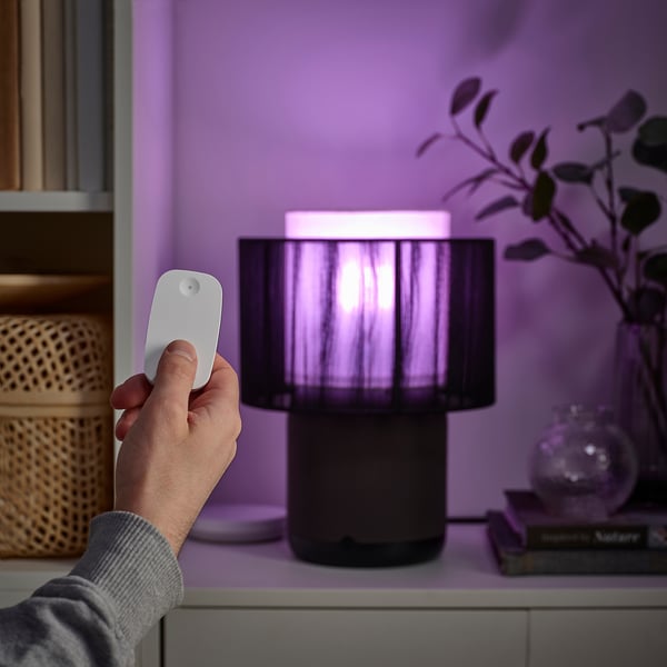 A hand holds a remote, controlling a stylish lamp with a cylindrical lampshade, emitting purple light.