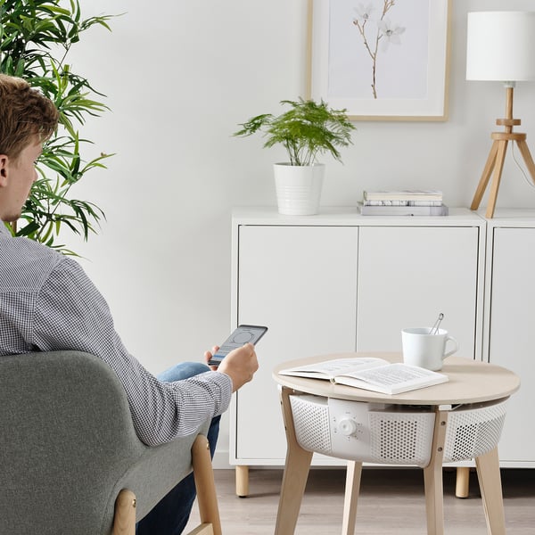 Person sitting on chair using smartphone, next to a table with air purifier, cup, and book.