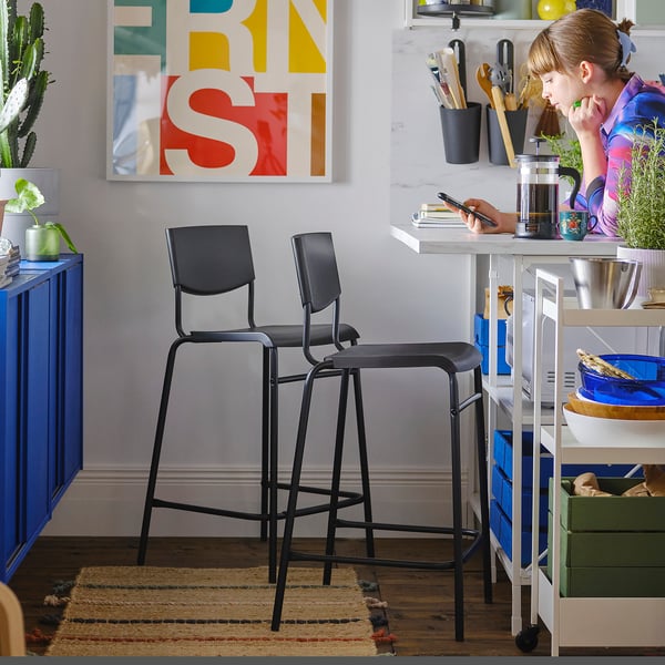Two black STIG bar stools near a counter where a person sits with a colourful shirt, surrounded by kitchen items and vibrant decor.