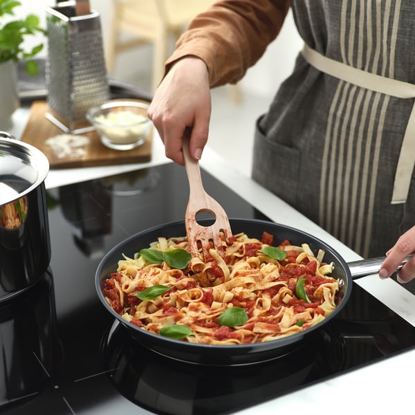 A person uses a wooden spaghetti server with a central hole to measure portions and grab spaghetti from a skillet.