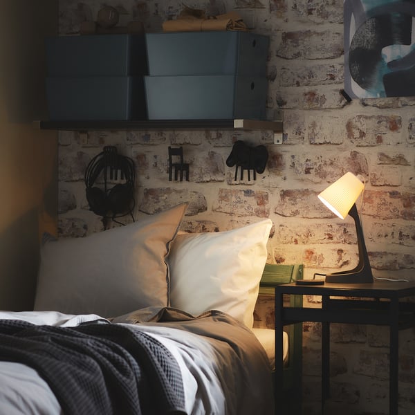 Bedroom with brick wall, white pillows, grey blanket, small black desk, lamp, and wall-mounted cabinets.