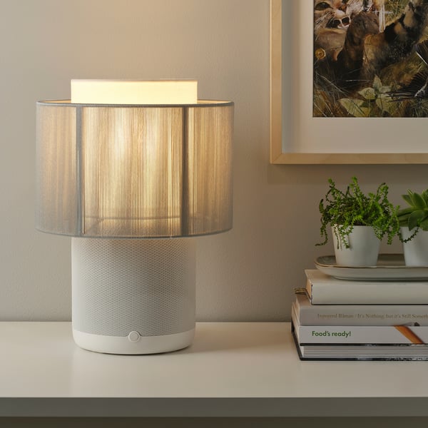 A modern lamp with a built-in wifi speaker on a table, playing light and music. Beside books and plants.