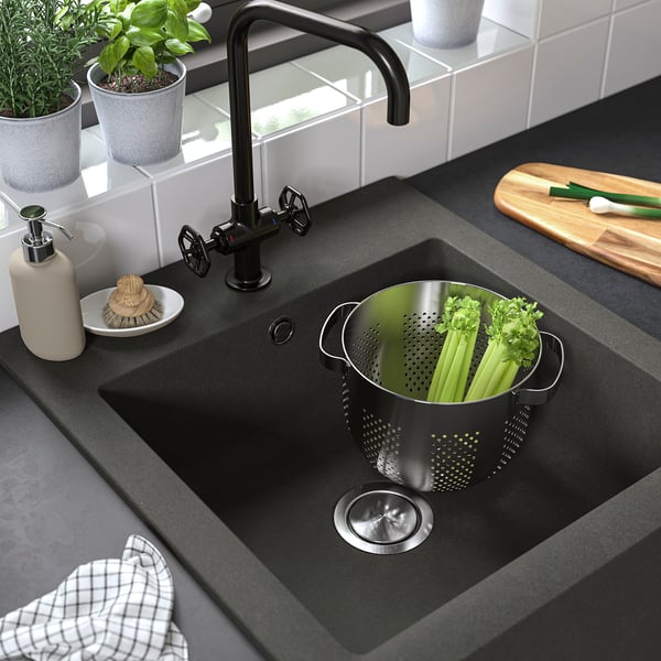 Black farmhouse sink with stainless steel strainer basket filled with celery. Modern kitchen setup with black tap and various accessories.