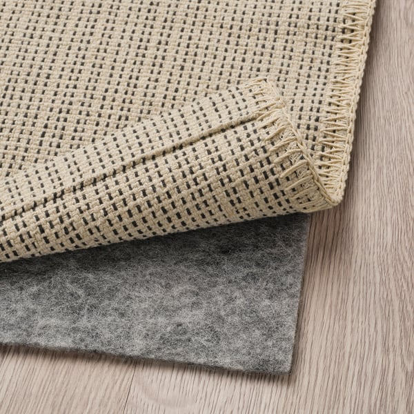 A beige woven rug with dark accents, placed on a grey non-slip pad on a wooden floor.