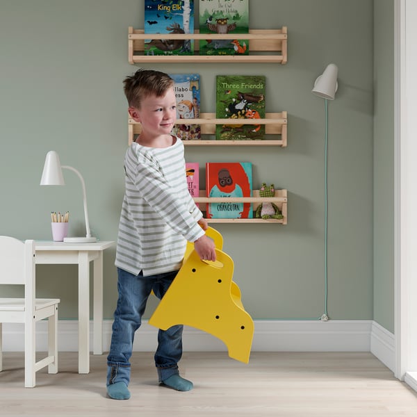 A young boy holding a bright yellow TROGEN stool in a room with wooden bookshelves and a white desk.