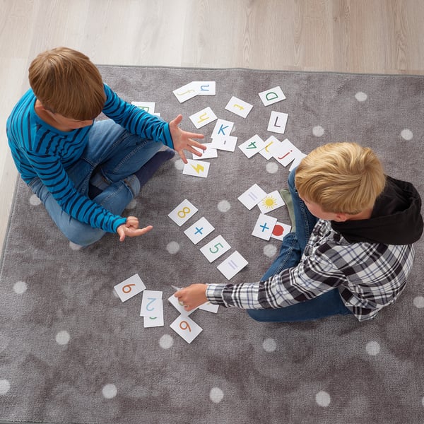 Two kids play with alphabet and number cards on a rug. One reaches for a card, the other observes.