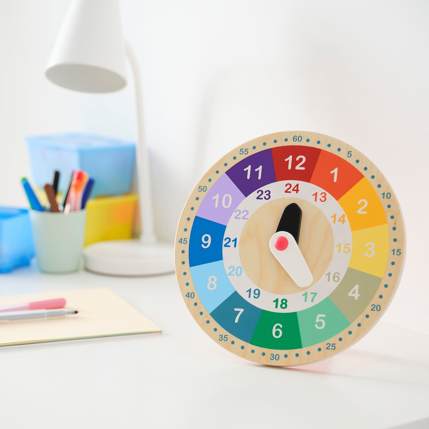 Colourful wood clock UNDERHÅLLA on desk with pencil holder, lamp, papers, time 9:00.