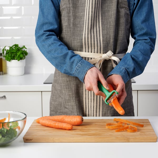 Person peeling carrots using UPPFYLLD peeler on wooden cutting board. Bowl with greens nearby.