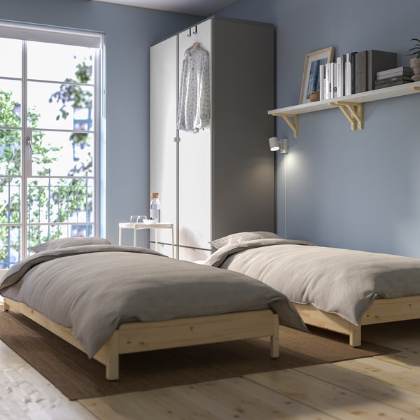 Two wooden platform beds, stacked as a sofa, beside a window with natural light. Grey bedding on both, simple modern decor.