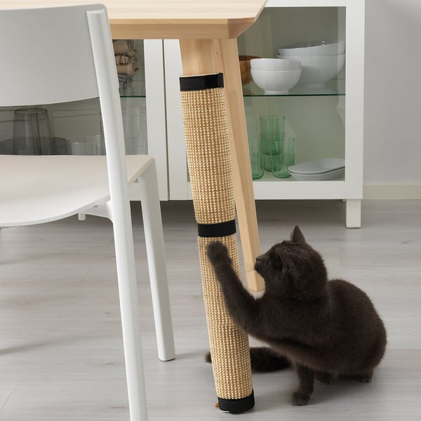 Black cat plays with beige and black UTSÅDD cat scratching mat wrapped around table leg in modern kitchen.