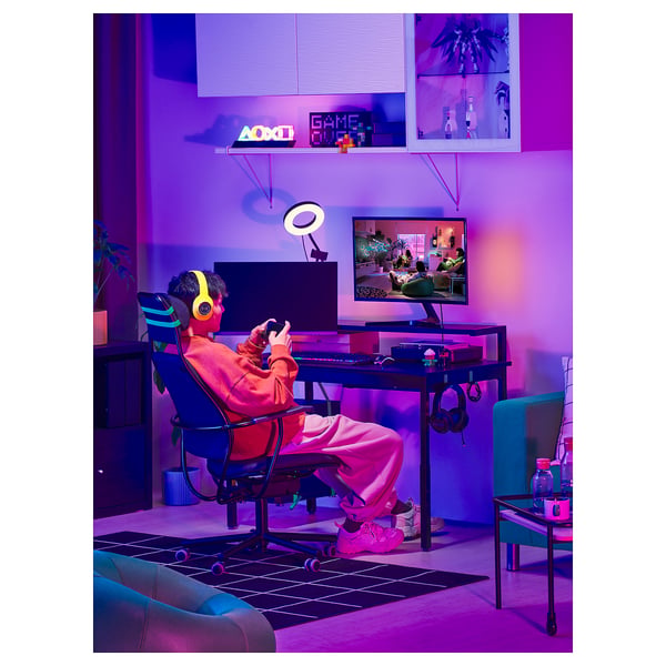 In a neon-lit room, a person plays a video game at a height-adjustable desk with a monitor, keyboard, and yellow headphones.