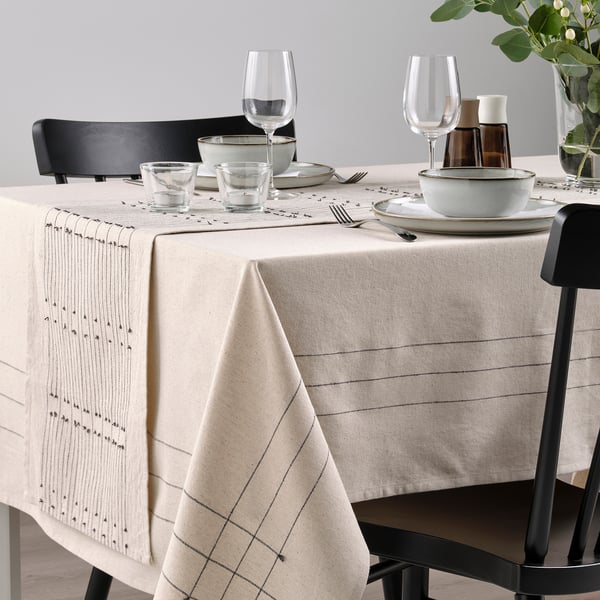 Dining table with beige runner, subtle patterns, tassels, black chairs, glassware, ceramic dishes for cosy meal.
