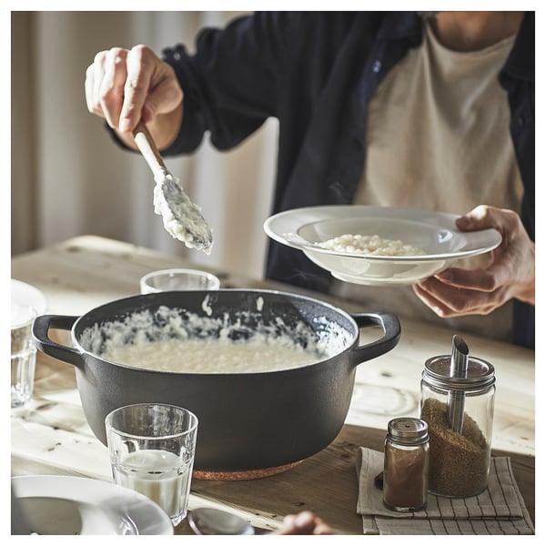 Person serving creamy rice from black VARDAGEN pan onto white plate on wooden table with seasonings and glasses.