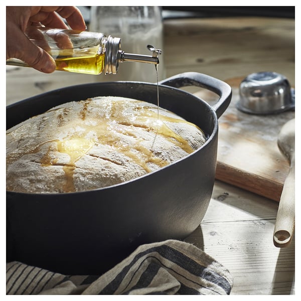 A black cast iron casserole has oil being poured on bread, on a wooden table.