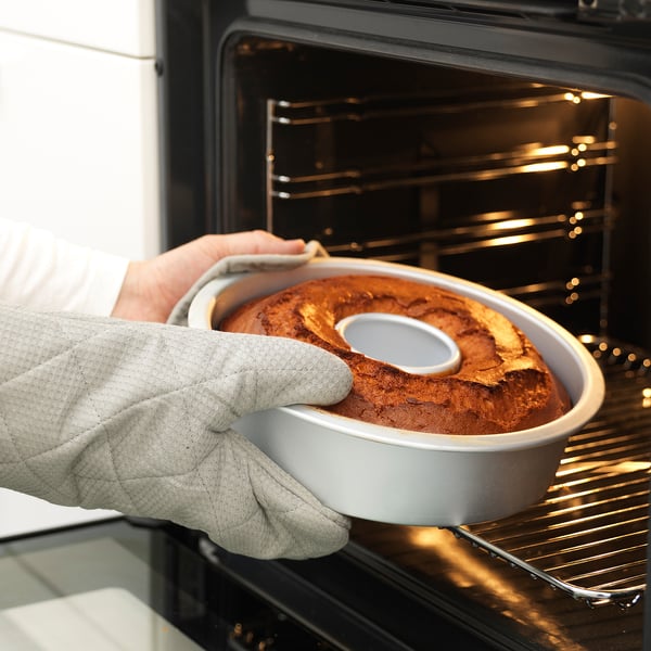 A person wearing oven gloves carefully removes a golden-brown bundt cake from an oven using a baking tin with a removable base.