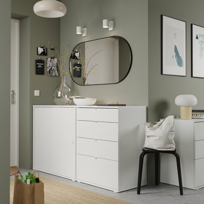 Modern green room with white furniture including a dresser, chair, and stool. Artwork, a mirror, and decorative items.