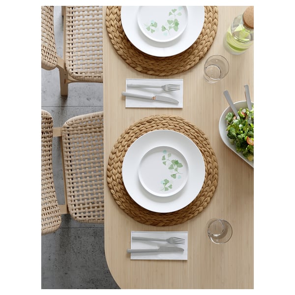 Two places set with white plates, cutlery, glasses, and a salad bowl on a light wooden table surrounded by wicker chairs.