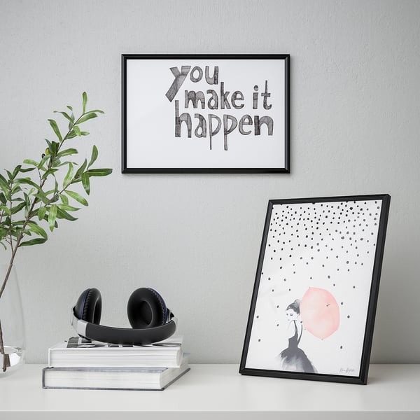 Framed posters on a desk. One says you make it happen, the other shows a person with a red dot. Headphones on books, plant beside.