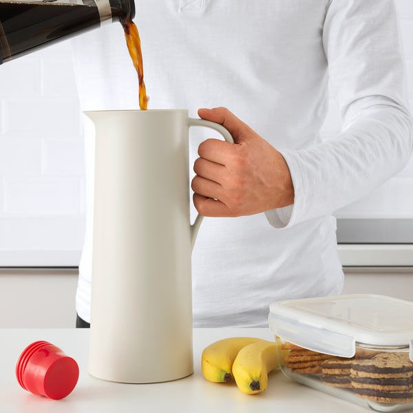 Person pouring coffee into a white BEHÖVD thermal flask on a white table with snacks.