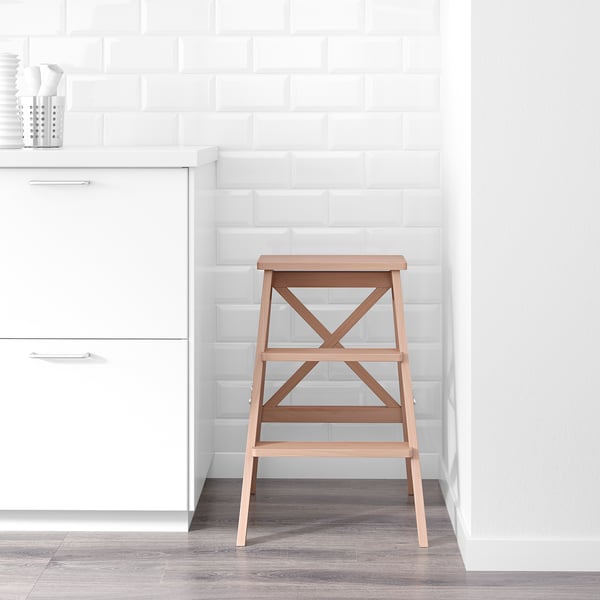 Wooden step ladder against white wall, next to kitchen cabinets. Folds for space-saving, sturdy, holds up to 100 kg.