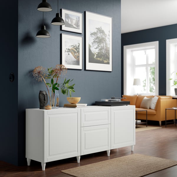 White BESTÅ sideboard with black pendant lights, and artwork set against dark blue wall beside entrance, near orange seating area in well-lit room.