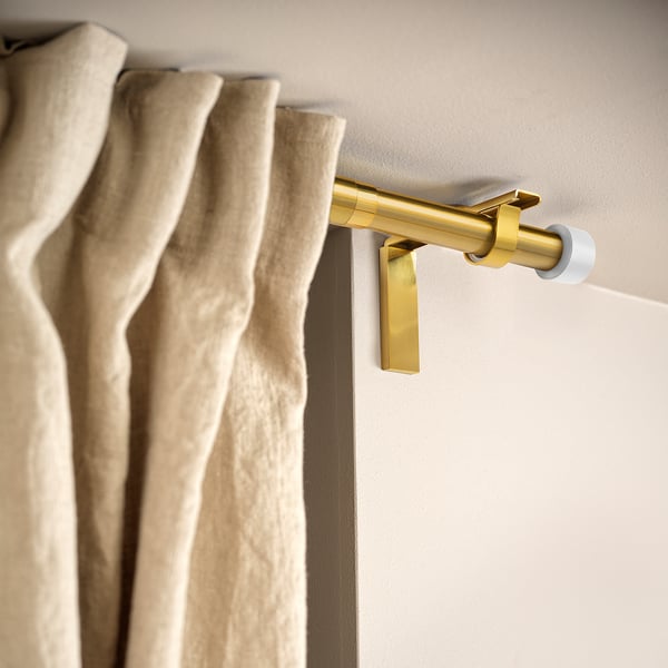 Golden curtain rod holder mounted on wall, holding beige curtains. Allows curtain placement near or far from window.