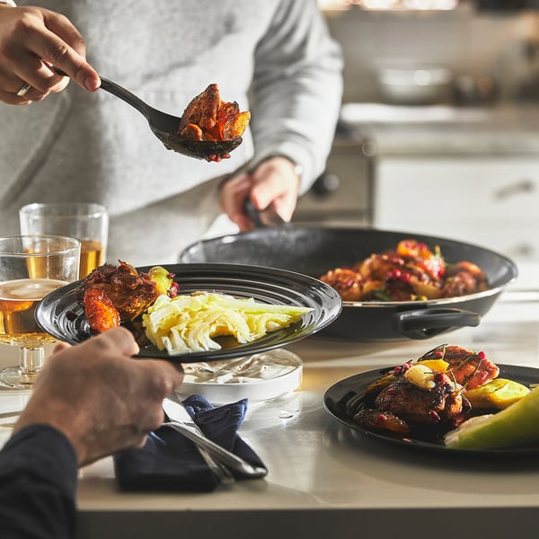 Two people prepare a meal with grilled chicken and side dishes in a large frying pan.