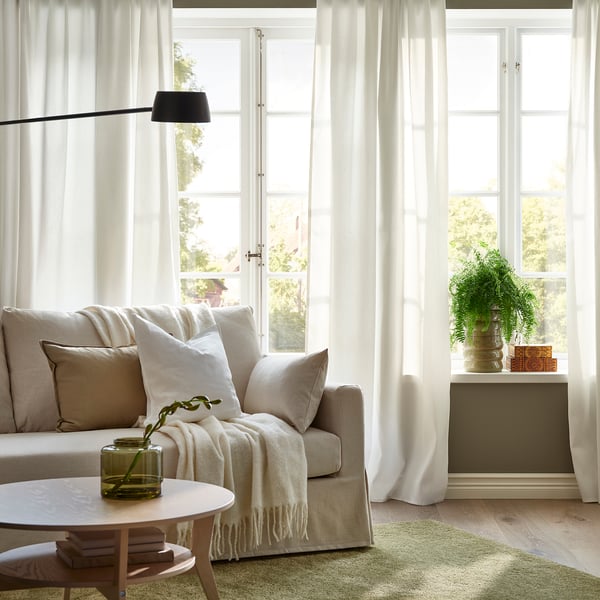 Cosy living room with beige sofa, white curtains, coffee table with plants and blanket, side table plant.