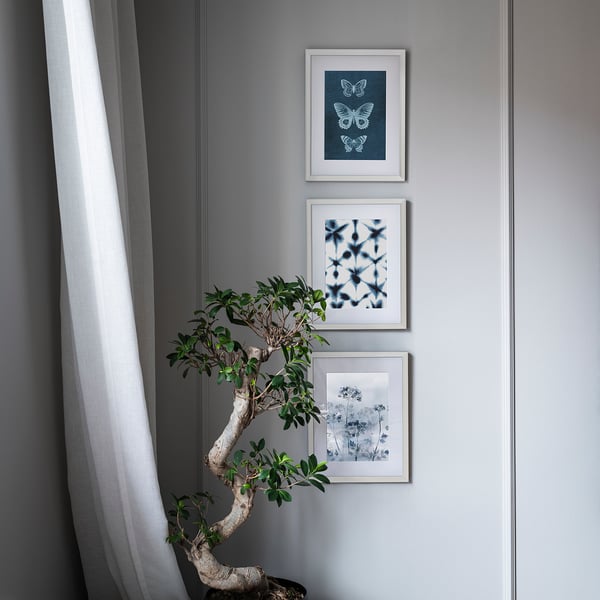 Three framed blue artworks hung vertically on grey wall beside a white curtain. Bottom artwork depicts plants in vase.