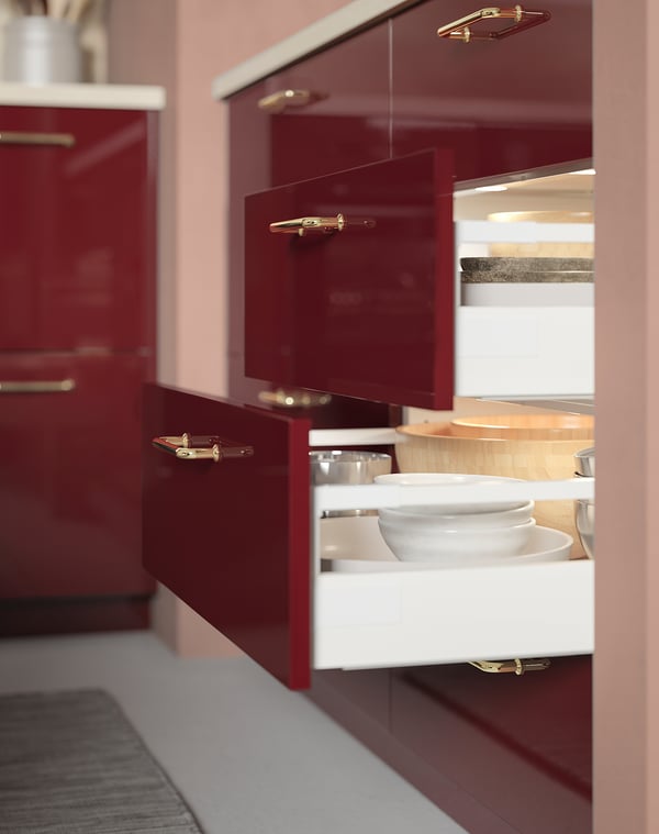 Red glossy kitchen cabinets with open drawers revealing white plates. Gold handles and led light inside drawers.