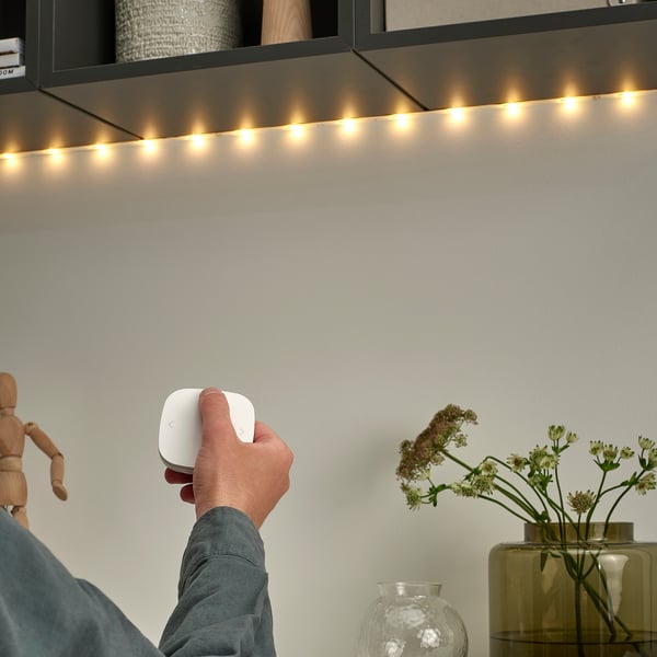 Hand holds ikea smart light controller; shelf has warm string lights; vase with flowers, wooden figure nearby.