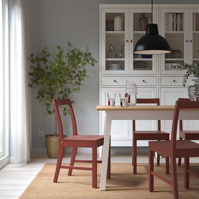 Red wooden PINNTORP chair with slanted back, around dining table on rug, white cabinet with glassware, plant. Cosy, minimalist, rustic-modern.
