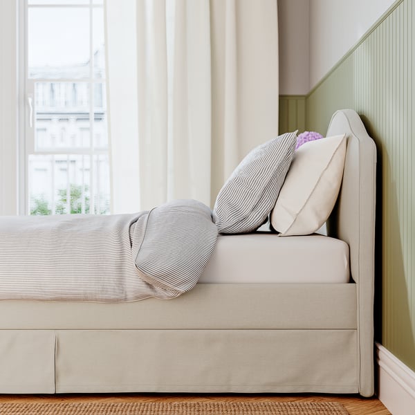 Beige bed with curved headboard, two pillows, striped blanket, near window with light curtains.