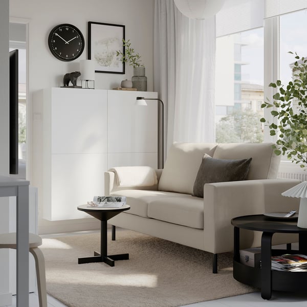 Modern living room with white SALTSJÖBADEN sofa, black side tables, and a large window. Bright, light walls and beige carpet create a cosy atmosphere.