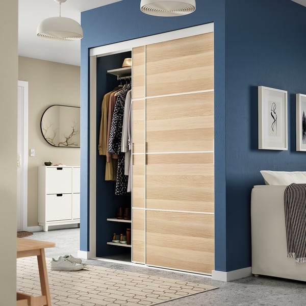SKYTTA sliding closet doors: light wood and glass panels. Tall, spans floor to ceiling, flanked by dark blue walls. Modern minimalist design.
