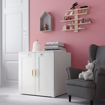 Kids room with pink walls, grey armchair, white cabinet, toy shelves, light-colored lamp, books, and soft toy.