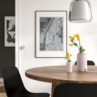 Modern dining table with wooden top, black chairs, and pendant light. Pink striped stoneware vases with yellow flowers. Urban nyc poster behind.