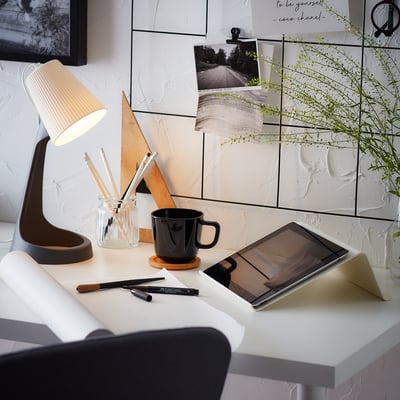 A minimalist workspace featuring a white desk with a lamp, tablet, and mug. A mirror with photos and plants decorates the wall.