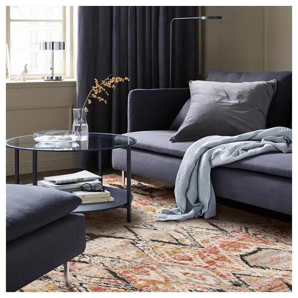 Modern living room with blue couch, glass coffee table, books, grey blanket, and multicoloured geometric rug.
