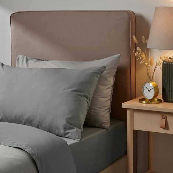 A bedding setup with an ULLVIDE brown pillowcase and grey sheets on a bed with a wooden headboard and a bedside table with a lamp and clock.