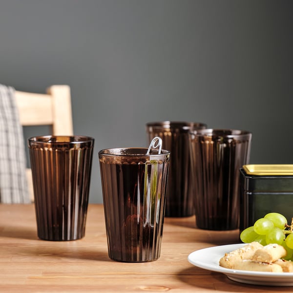 Six dark, ribbed glasses on a wooden table, one with a spoon, next to a plate of cookies and grapes.