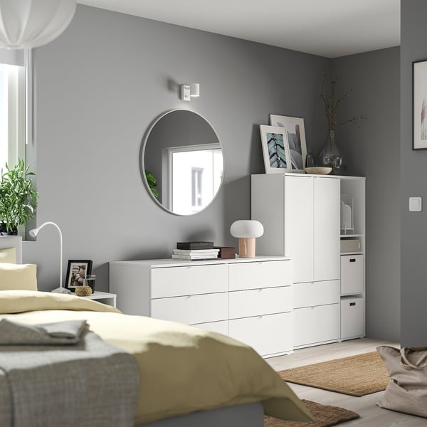 Modern bedroom with grey walls, white furniture, yellow blanket, round mirror, lamp, and decorative items.