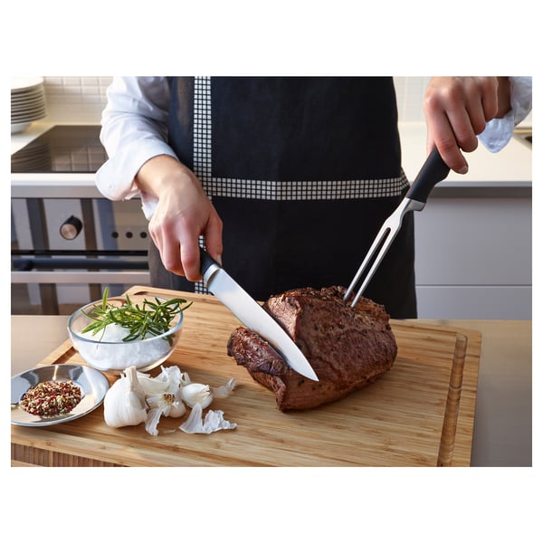 Person carving roast with VÖRDA knife and fork; ingredients and kitchen tools on wooden board.