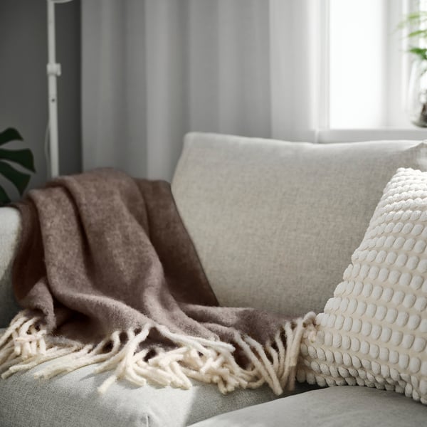 ALLFRAKEN: taupe blanket, 100% wool, covers light beige sofa, large pillow with white pattern, by window, cosy setup.