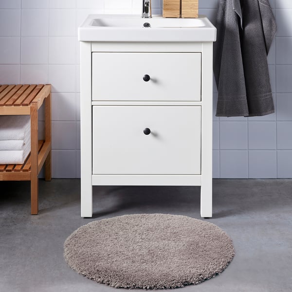A white vanity with a sink and drawers, featuring a plush, beige ALMTJÄRN bath mat and a wooden bench with towels and a basket.