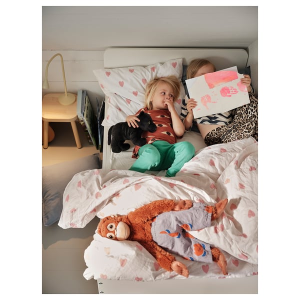 Two children on a bed with heart-patterned duvet, one reading, one drawing. Stuffed animals nearby.