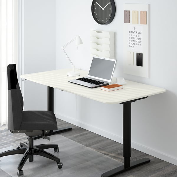 Modern home office with white BEKANT desk, grey chair, laptop, lamp, clock, and calendar.