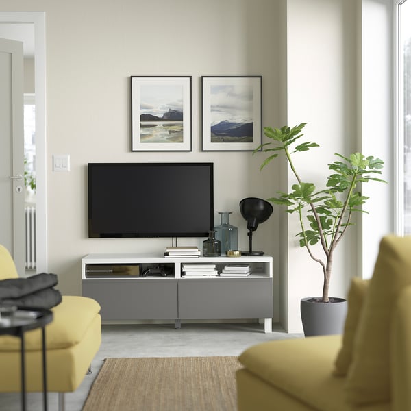 Modern living room with BESTÅ tv bench in grey, featuring flat-screen tv, yellow chairs, large plant, and framed artwork.