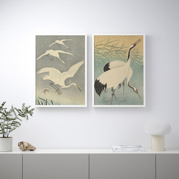 Two framed crane art prints hang on a wall above a white cabinet. A potted plant and a white lamp are on the cabinet.